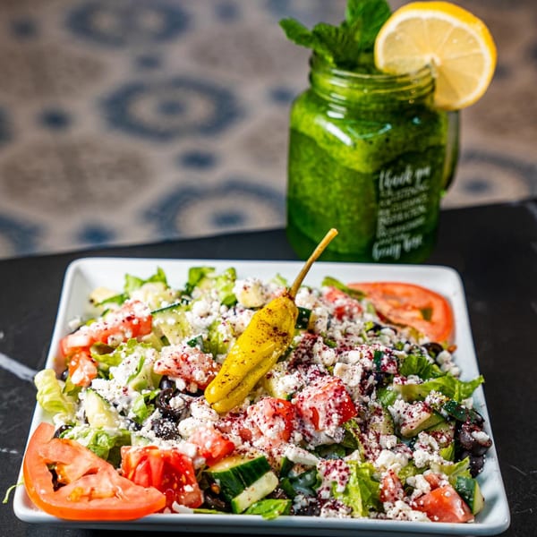 A fresh Mediterranean salad with feta, tomatoes, olives, and a pepper, alongside a green drink.